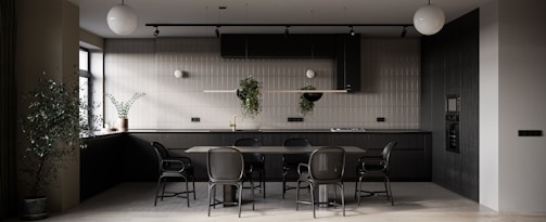 A modern kitchen primarily featuring dark tones with a minimalistic design. There is a large dining table with matching chairs in the center. The kitchen counter is equipped with modern appliances, and potted plants are hanging from a rail as well as placed on the counter, adding a touch of greenery. The ambient lighting includes spherical ceiling lamps, and natural light is streaming in through a window on the left side.