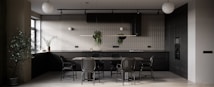A modern kitchen primarily featuring dark tones with a minimalistic design. There is a large dining table with matching chairs in the center. The kitchen counter is equipped with modern appliances, and potted plants are hanging from a rail as well as placed on the counter, adding a touch of greenery. The ambient lighting includes spherical ceiling lamps, and natural light is streaming in through a window on the left side.