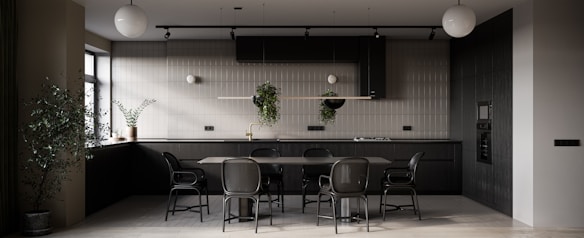 A modern kitchen primarily featuring dark tones with a minimalistic design. There is a large dining table with matching chairs in the center. The kitchen counter is equipped with modern appliances, and potted plants are hanging from a rail as well as placed on the counter, adding a touch of greenery. The ambient lighting includes spherical ceiling lamps, and natural light is streaming in through a window on the left side.