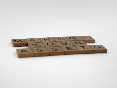 Wooden Scrabble tiles arranged in an organized pattern spell out the phrase 'MANAGE YOUR ASSETS' on a plain white background.