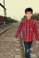 A child wearing a red and black plaid shirt and jeans walks down a railway track. The background features railway tracks extending into the distance, surrounded by greenery and fences.
