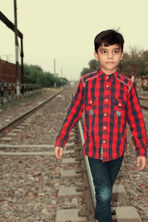 A child wearing a red and black plaid shirt and jeans walks down a railway track. The background features railway tracks extending into the distance, surrounded by greenery and fences.