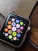 A smartwatch with a black screen displaying colorful app icons arranged in a grid. The watch has a metallic, gold-colored mesh strap and is resting on a wooden surface. The app icons include a mail icon, a music note, a heart symbol, and fitness-related symbols, among others.