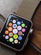 A smartwatch with a black screen displaying colorful app icons arranged in a grid. The watch has a metallic, gold-colored mesh strap and is resting on a wooden surface. The app icons include a mail icon, a music note, a heart symbol, and fitness-related symbols, among others.