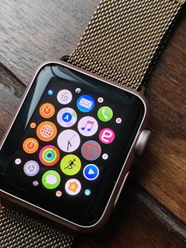A smartwatch with a black screen displaying colorful app icons arranged in a grid. The watch has a metallic, gold-colored mesh strap and is resting on a wooden surface. The app icons include a mail icon, a music note, a heart symbol, and fitness-related symbols, among others.