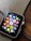 A smartwatch with a black screen displaying colorful app icons arranged in a grid. The watch has a metallic, gold-colored mesh strap and is resting on a wooden surface. The app icons include a mail icon, a music note, a heart symbol, and fitness-related symbols, among others.