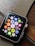A smartwatch with a black screen displaying colorful app icons arranged in a grid. The watch has a metallic, gold-colored mesh strap and is resting on a wooden surface. The app icons include a mail icon, a music note, a heart symbol, and fitness-related symbols, among others.