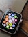 A smartwatch with a black screen displaying colorful app icons arranged in a grid. The watch has a metallic, gold-colored mesh strap and is resting on a wooden surface. The app icons include a mail icon, a music note, a heart symbol, and fitness-related symbols, among others.