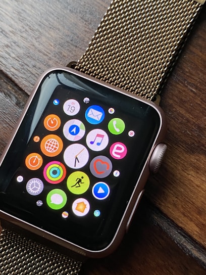 A smartwatch with a black screen displaying colorful app icons arranged in a grid. The watch has a metallic, gold-colored mesh strap and is resting on a wooden surface. The app icons include a mail icon, a music note, a heart symbol, and fitness-related symbols, among others.