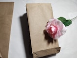 A stack of brown paper bags is placed on a white surface with a pink artificial rose resting on top of the stack. The bags are neatly folded and aligned, creating a clean and organized look.