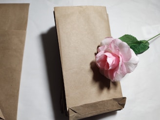 A stack of brown paper bags is placed on a white surface with a pink artificial rose resting on top of the stack. The bags are neatly folded and aligned, creating a clean and organized look.
