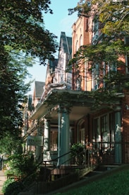 A row of historical brick buildings with detailed architectural features is partially covered by lush green trees. The buildings have tall windows, ornate columns, and intricate cornices. A sidewalk is lined with plants, and a sign saying 'STUDIO' is visible, suggesting a commercial or artistic space in one of the houses.