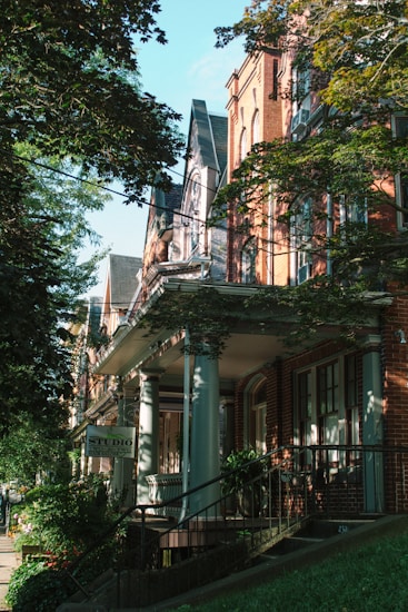 A row of historical brick buildings with detailed architectural features is partially covered by lush green trees. The buildings have tall windows, ornate columns, and intricate cornices. A sidewalk is lined with plants, and a sign saying 'STUDIO' is visible, suggesting a commercial or artistic space in one of the houses.