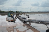 A small excavator is operating in a salt flat, moving salt onto a conveyor belt. The background features industrial structures, a line of trees, and a distant cityscape under a cloudy sky.