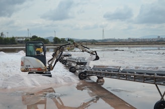 A small excavator is operating in a salt flat, moving salt onto a conveyor belt. The background features industrial structures, a line of trees, and a distant cityscape under a cloudy sky.