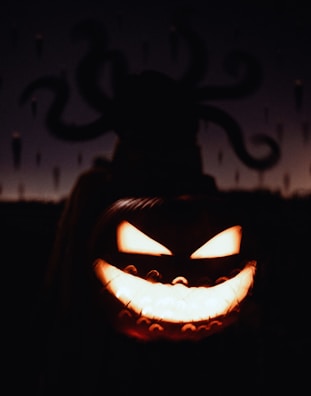A carved pumpkin with an eerie, menacing face is illuminated from within. In the background, a dark, shadowy figure with tentacle-like appendages is silhouetted against a dimly lit sky.
