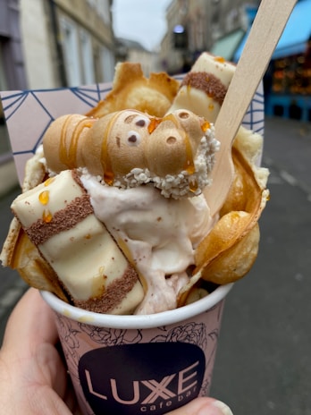 A decorative dessert in a cup from Luxe cafe bar, featuring bubble waffles, vanilla chocolate wafer rolls, and a dollop of ice cream, all topped with syrup and a wooden spoon. The background shows a blurred street scene.