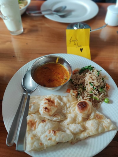A plate of food featuring a serving of naan bread, a bowl of lentil curry, and a portion of vegetable fried rice. The table setting includes a spoon and fork on the side of the plate. In the background, there is a glass with a white drink and a small note card with the text 'food is love' and a heart.