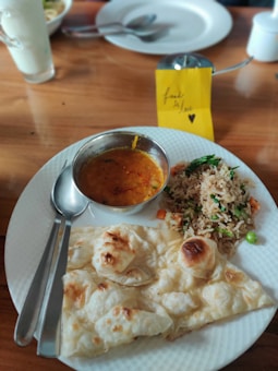 A plate of food featuring a serving of naan bread, a bowl of lentil curry, and a portion of vegetable fried rice. The table setting includes a spoon and fork on the side of the plate. In the background, there is a glass with a white drink and a small note card with the text 'food is love' and a heart.