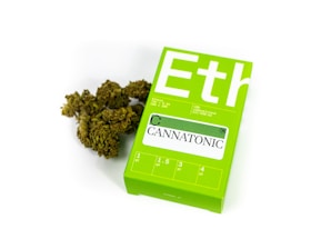 A green package labeled 'Cannatonic' is lying next to a small pile of green botanical material resembling dried cannabis. The package contains dosage information and is designed with clear, bold lettering on a bright green background.