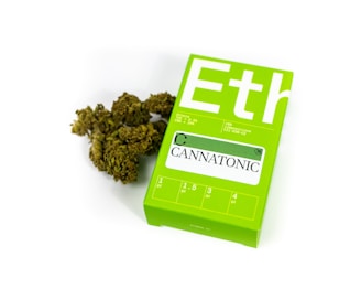 A green package labeled 'Cannatonic' is lying next to a small pile of green botanical material resembling dried cannabis. The package contains dosage information and is designed with clear, bold lettering on a bright green background.