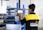 A person is interacting with a digital control panel in an industrial setting. The individual is wearing a black and yellow jacket with text on the back. The background features metal shelves and equipment, indicating a workshop or factory environment.