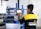 A person is interacting with a digital control panel in an industrial setting. The individual is wearing a black and yellow jacket with text on the back. The background features metal shelves and equipment, indicating a workshop or factory environment.