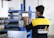 A person is interacting with a digital control panel in an industrial setting. The individual is wearing a black and yellow jacket with text on the back. The background features metal shelves and equipment, indicating a workshop or factory environment.