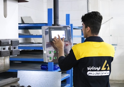 A person is interacting with a digital control panel in an industrial setting. The individual is wearing a black and yellow jacket with text on the back. The background features metal shelves and equipment, indicating a workshop or factory environment.