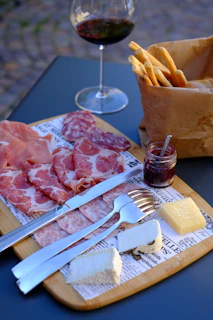 A wooden serving board features an assortment of cured meats, including slices of prosciutto and salami, placed alongside pieces of white and yellow cheese. A small jar of jam with a spoon is positioned on the board next to a knife and fork. A brown paper bag is filled with breadsticks, and a glass of red wine is positioned in the background.