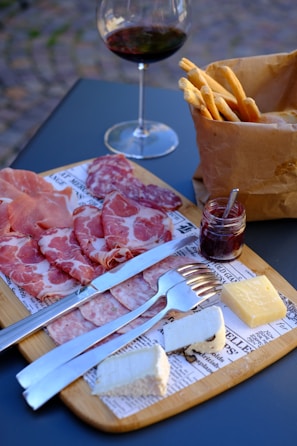 A wooden serving board features an assortment of cured meats, including slices of prosciutto and salami, placed alongside pieces of white and yellow cheese. A small jar of jam with a spoon is positioned on the board next to a knife and fork. A brown paper bag is filled with breadsticks, and a glass of red wine is positioned in the background.