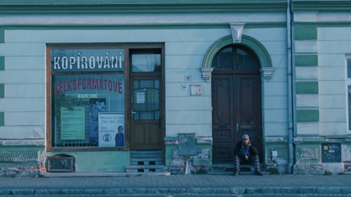A storefront with a large window displaying the word 'Kopírování' and other signage in a foreign language. The building has a pale green facade with dark green accents and appears weathered. A person is sitting on the steps near a wooden door, wearing a hat and jacket, seemingly in a reflective or waiting position.