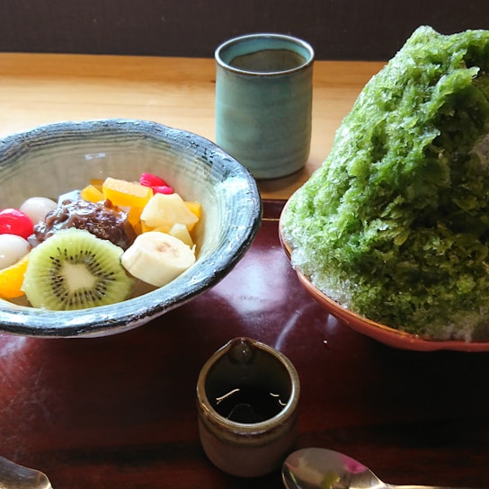 A traditional Japanese dessert setup featuring a bowl with various fruits including kiwi, banana, and cherries, alongside azuki beans and mochi. Next to it is a large serving of green shaved ice in a bowl, with a small pitcher of syrup, a cup, and a spoon on a wooden tray.