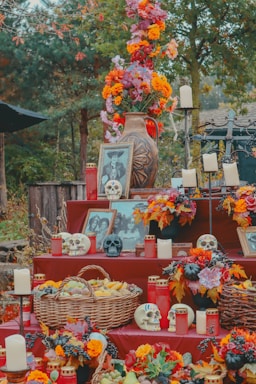 An ornate altar is adorned with vibrant orange and purple flowers, candlesticks, red candles, and intricately decorated skulls. Framed photographs and a large, patterned vase contribute to the elaborate setup. A wicker basket filled with fruits and surrounded by autumn leaves enhances the festive and commemorative display.