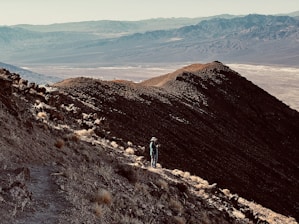 A lone hiker stands on a rocky mountain trail, overlooking a vast, arid landscape with distant mountain ranges under a clear sky.