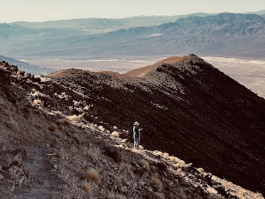 A lone hiker stands on a rocky mountain trail, overlooking a vast, arid landscape with distant mountain ranges under a clear sky.