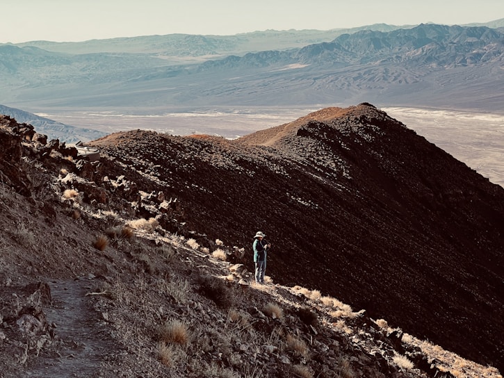 A hiker pausing on a rocky trail with expansive mountain views under a clear blue sky.