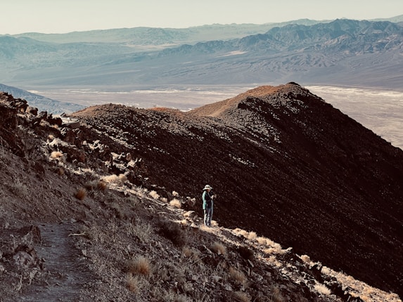 A lone hiker stands on a rocky mountain trail, overlooking a vast, arid landscape with distant mountain ranges under a clear sky.