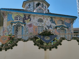 A vibrant and intricately decorated house featuring colorful mosaics and paintings of butterflies and various abstract designs. The facade includes multiple windows with ornate iron grills and a prominently tiled roof with blue accents. Flowers and greenery are arranged along the balcony, adding to the lively appearance.