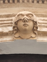 A detailed sculpture of a human face with stylized hair carved into a stone archway, featuring a serene expression. The craftsmanship showcases intricate details and symmetry.