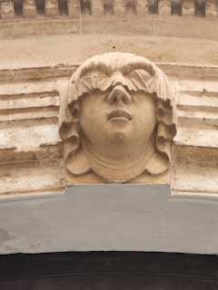 A detailed sculpture of a human face with stylized hair carved into a stone archway, featuring a serene expression. The craftsmanship showcases intricate details and symmetry.