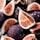 A close-up view of several figs, some of which are whole while others are cut open to reveal their intricate, seed-filled interiors. The figs are dark purple on the outside, with their inner flesh displaying a rich, reddish hue and numerous seeds. They appear fresh, with droplets of water visible on their surfaces.