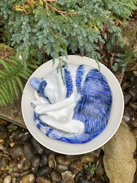A sculpture of a blue and white cat curled up in a circular dish, surrounded by greenery and pebbles. The sculpture is detailed, mimicking the appearance of a sleeping cat, with textured fur visible. The dish sits on a background of stones and fern leaves, adding a natural setting to the scene.
