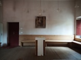 A simple, minimalist room with a central podium, a wooden bench along the walls, and a small religious painting on the wall. The room has muted colors and is dimly lit with hanging lights.