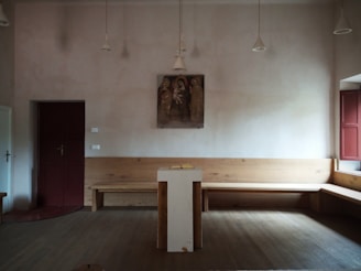 A simple, minimalist room with a central podium, a wooden bench along the walls, and a small religious painting on the wall. The room has muted colors and is dimly lit with hanging lights.