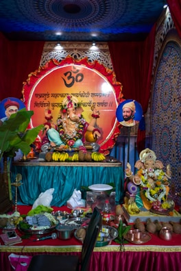 A vibrant display of a Hindu religious setup featuring a statue of Lord Ganesha adorned with colorful garlands of flowers and decorative elements. In the background, a large circular panel with Sanskrit script and a symbol is present. The arrangement includes offerings such as bananas and coconuts. The setting is ornate with rich colors and traditional patterns.