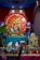 A vibrant display of a Hindu religious setup featuring a statue of Lord Ganesha adorned with colorful garlands of flowers and decorative elements. In the background, a large circular panel with Sanskrit script and a symbol is present. The arrangement includes offerings such as bananas and coconuts. The setting is ornate with rich colors and traditional patterns.