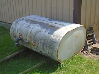 A large, cylindrical metal tank lies on its side on the ground next to a corrugated metal building. The tank appears rusted and weathered, with visible pipes attached to its side. It rests on wooden beams on the grass, with some miscellaneous items nearby.