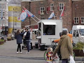 A street scene with a mobile food stand. People are gathered around the stand, engaging in conversation. The surrounding area includes historic buildings and trees, with colorful flags on the food stand. Pedestrians, including a child in a stroller, and a person with a bicycle, are visible. The scene has an urban, lively atmosphere.