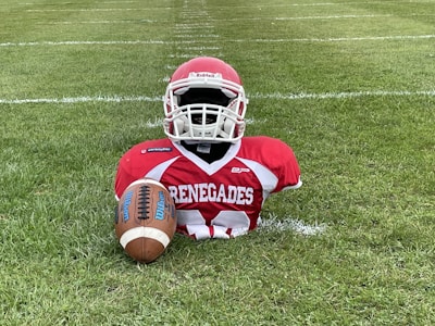 An American football dummy is placed on a field. It is wearing a red and white jersey with the word 'Renegades' printed on it, along with a red helmet. A brown Wilson football is positioned in front of the dummy.